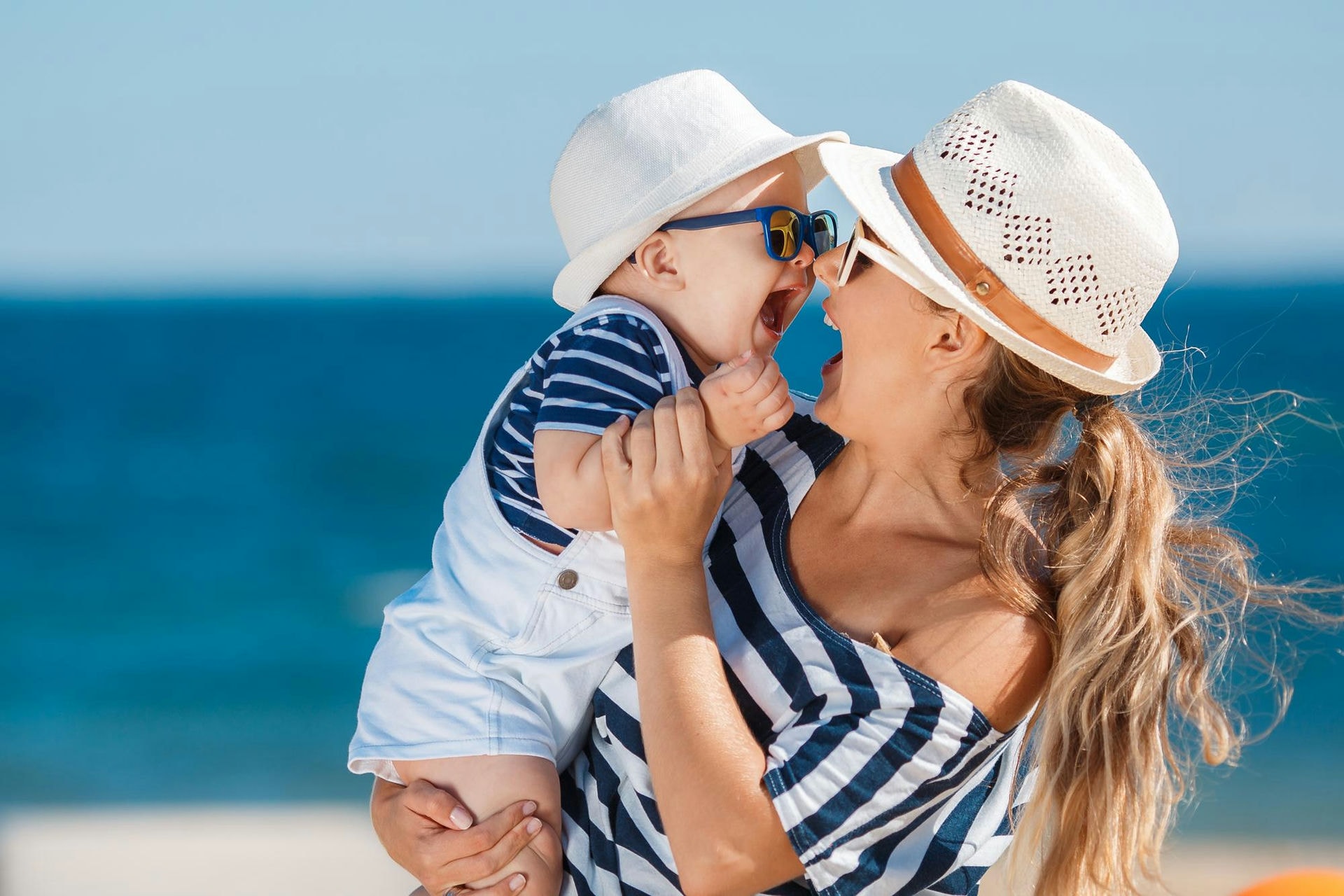 Kobieta w kapeluszu przytula roześmiane dziecko w okularach na tle morza i plaży