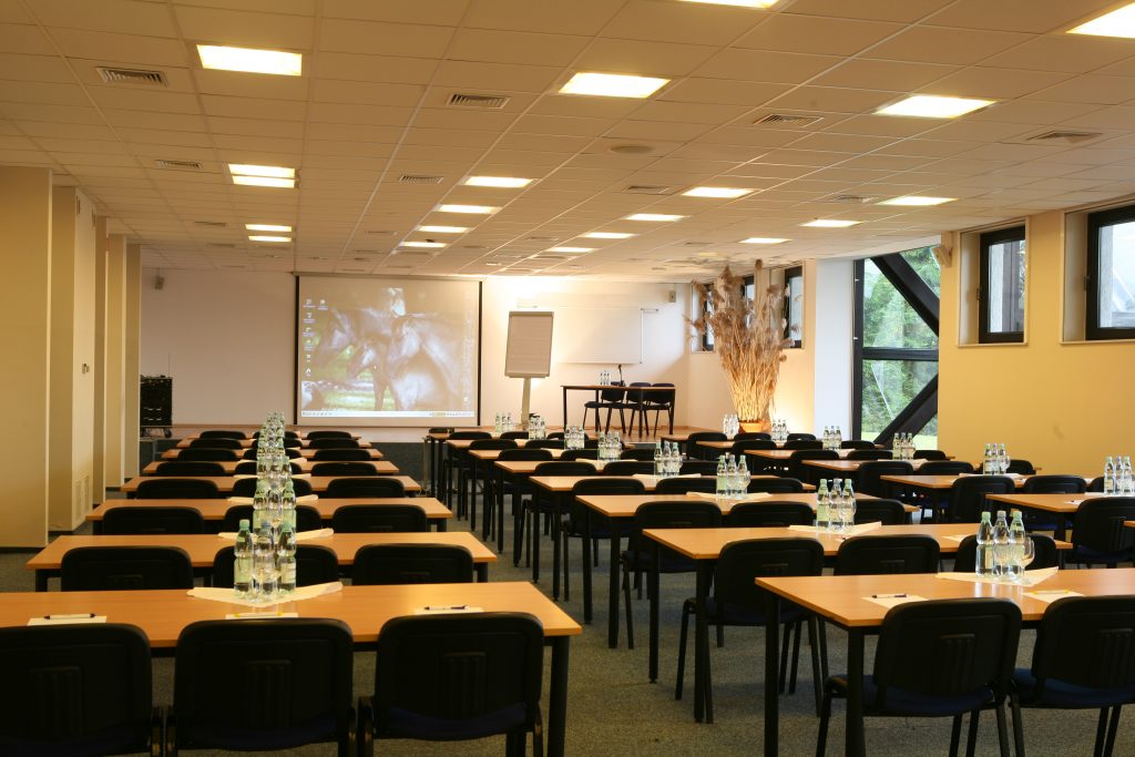 Sala konferencyjna z rzędami stołów i krzeseł, ekranem projekcyjnym, tablicą oraz wodą dla uczestników