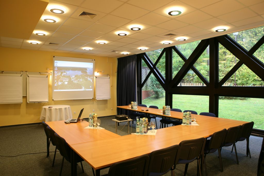 Sala konferencyjna ze stołami w podkowę, projektorem, tablicami flipchart i dużymi oknami wychodzącymi na ogród