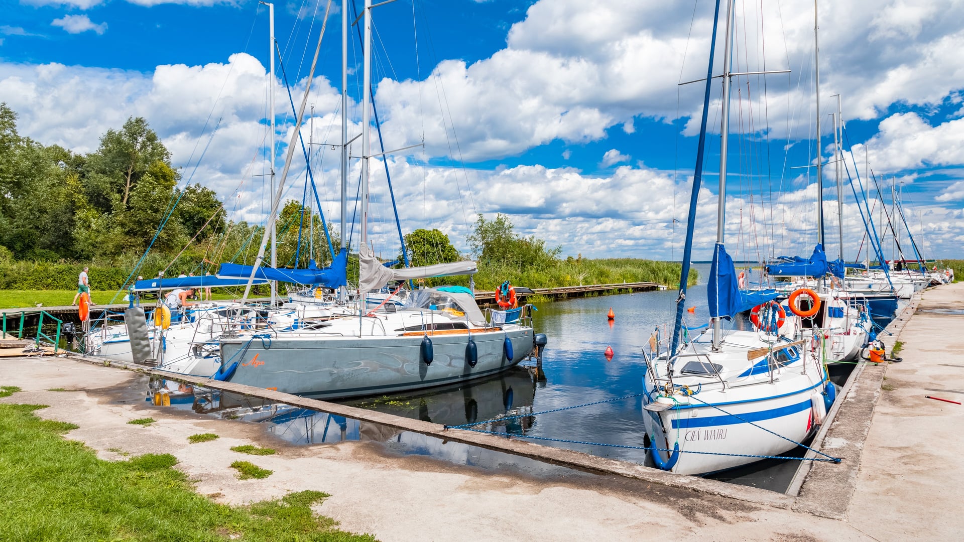 Żaglówki zacumowane w marinie nad jeziorem, z pomostami, zielenią i pochmurnym niebem w tle