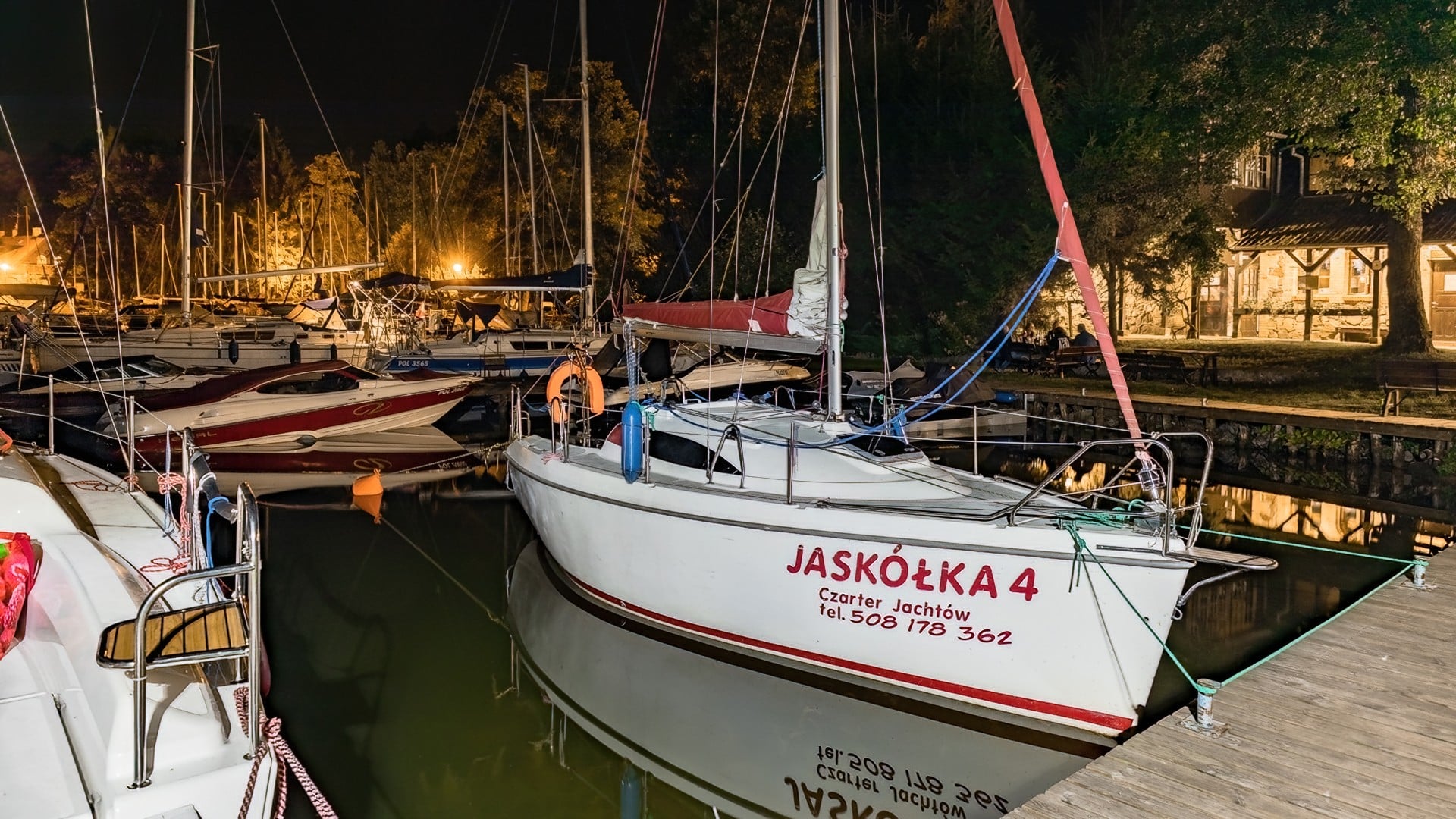 Nocna przystań żeglarska z licznymi jachtami; na pierwszym planie biały jacht „Jaskółka 4” przy pomoście