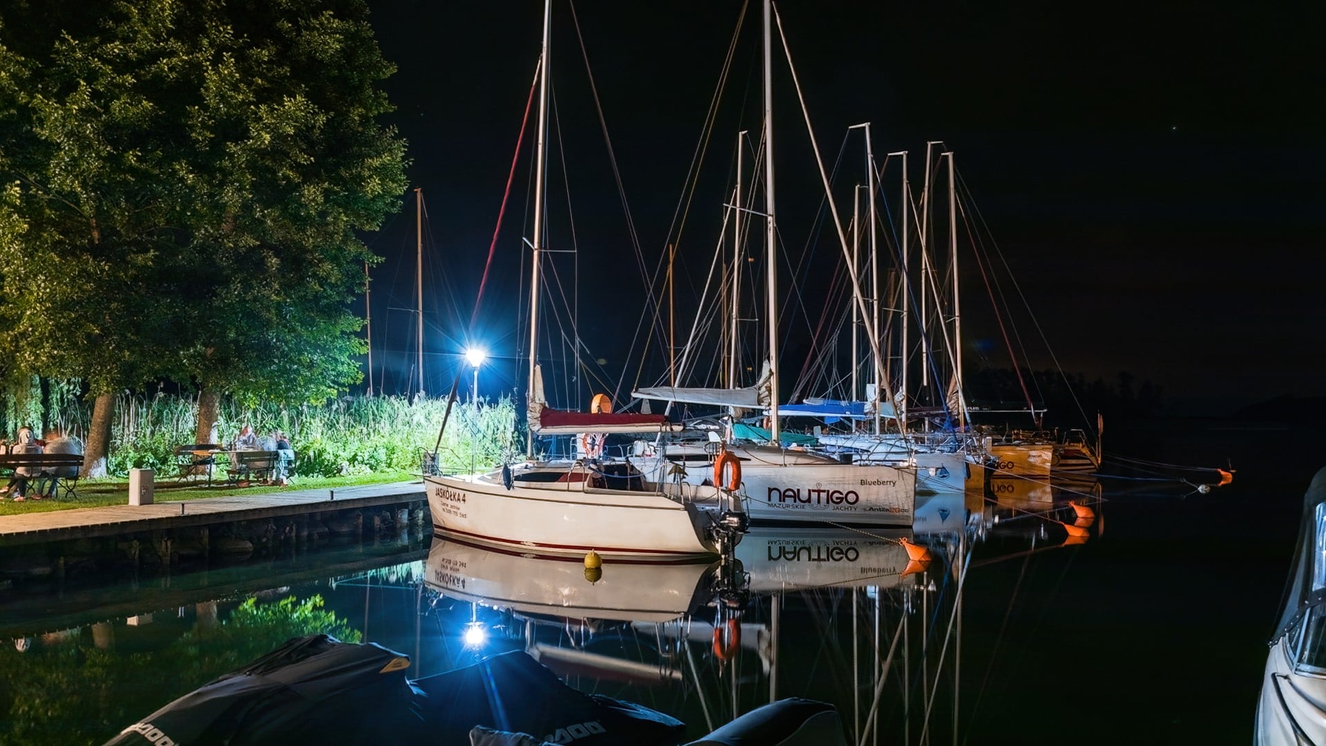 Oświetlona nocą przystań z żaglówkami odbijającymi się w spokojnej wodzie i drzewami na brzegu