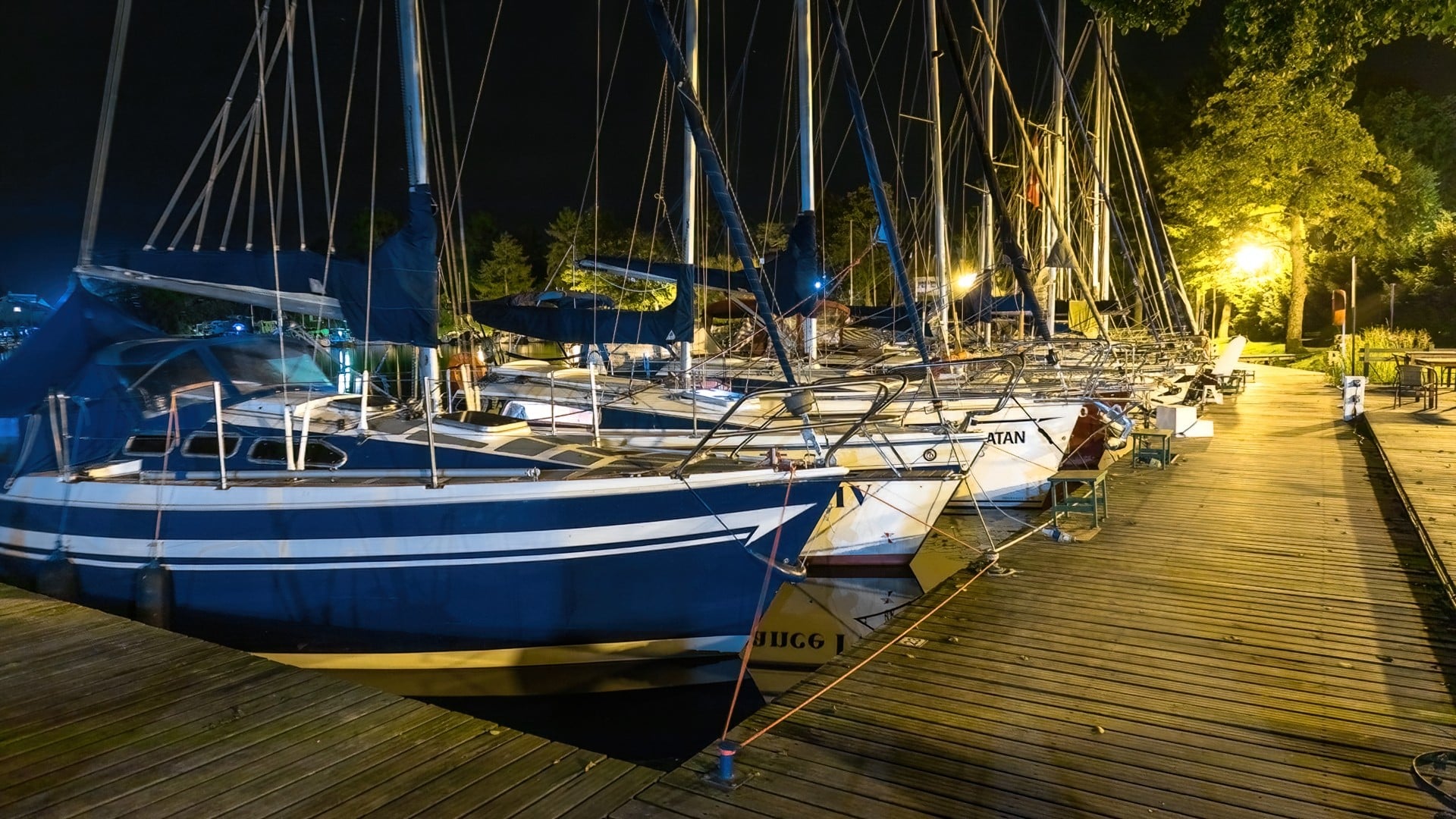 Oświetlona nocą marina z licznymi żaglówkami zacumowanymi przy drewnianym pomoście
