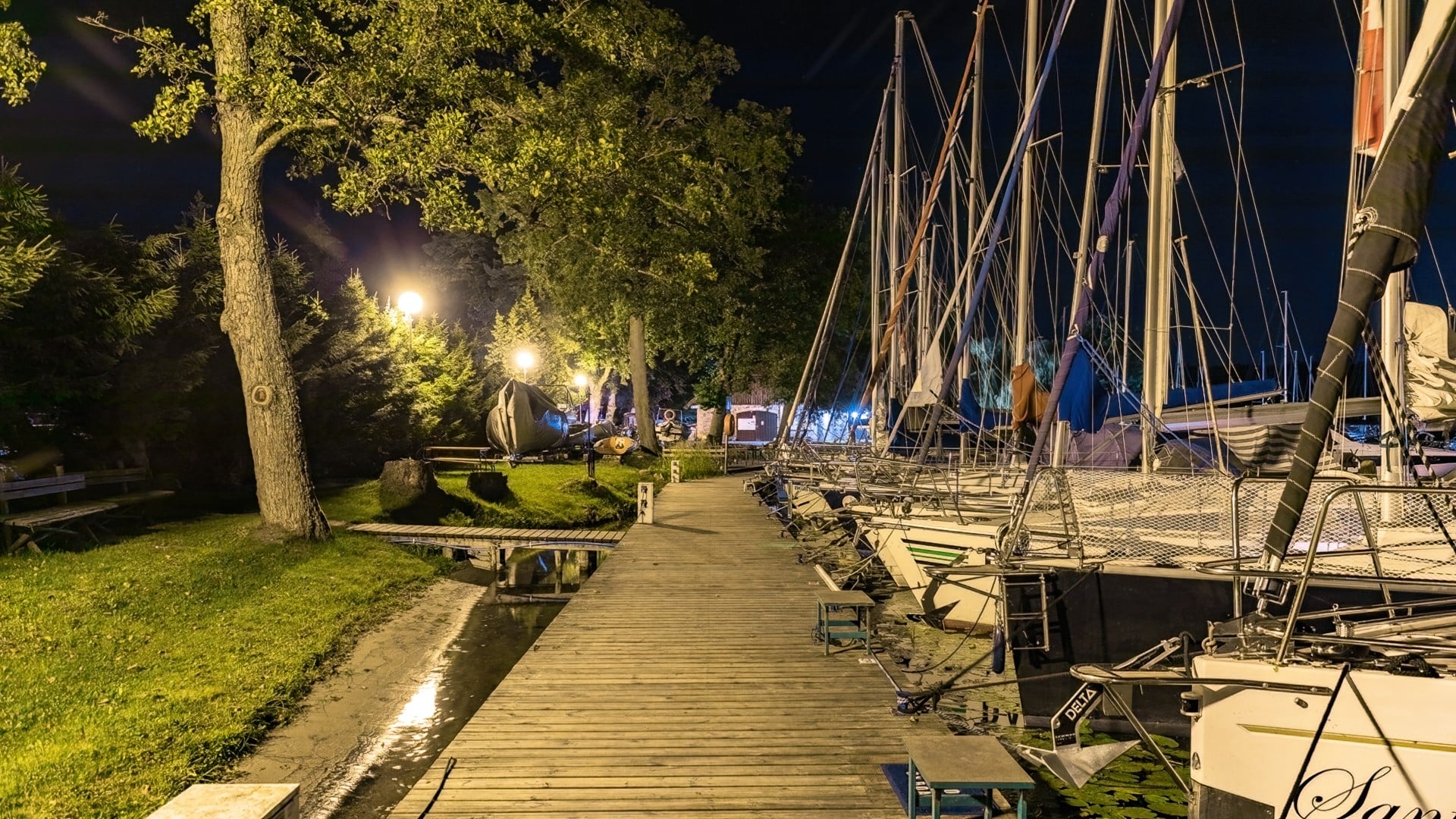 Nocą oświetlony pomost wśród drzew i licznych łodzi żaglowych zacumowanych przy brzegu jeziora