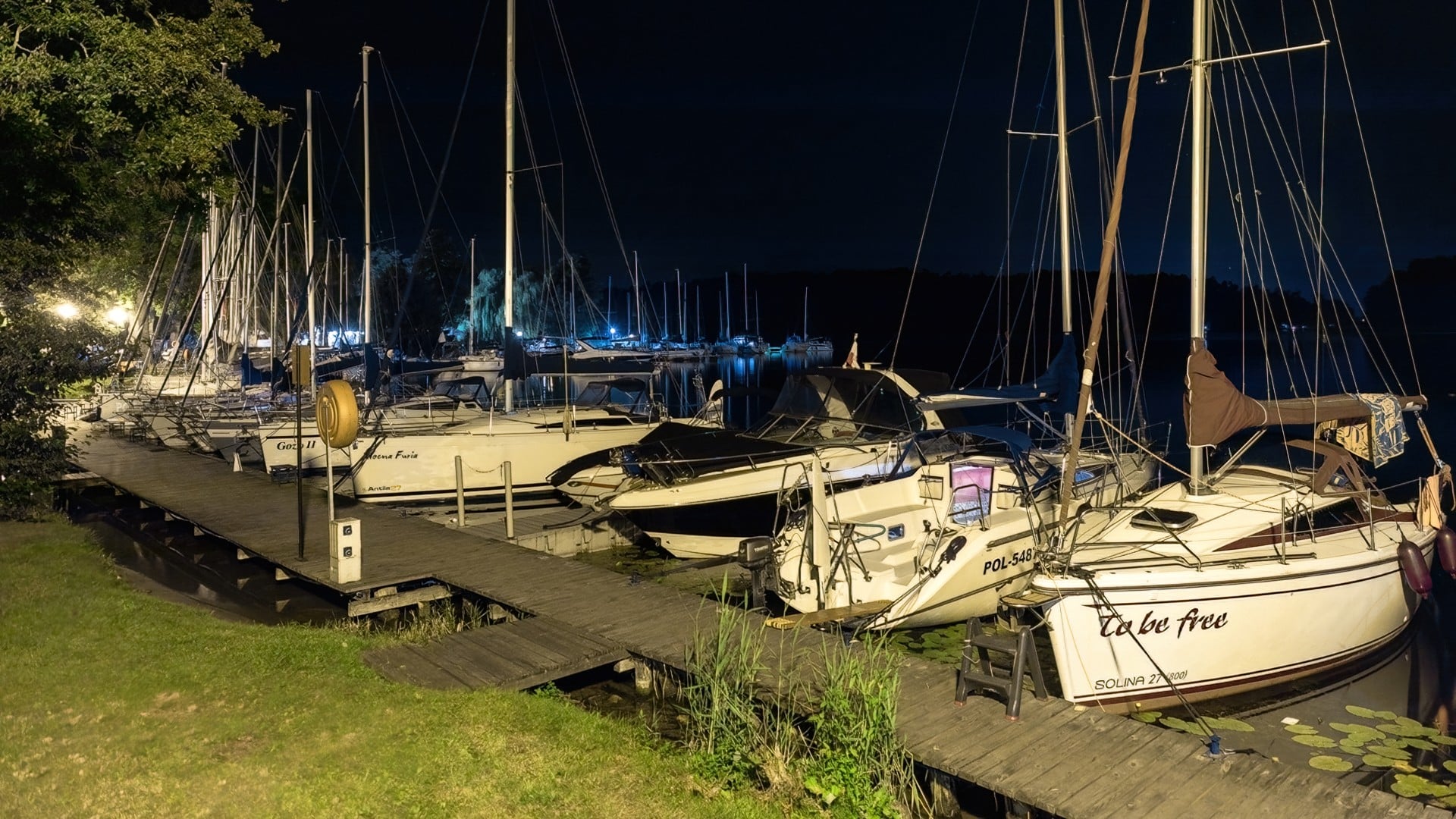 Oświetlona nocą przystań z licznymi żaglówkami zacumowanymi przy drewnianym pomoście nad jeziorem