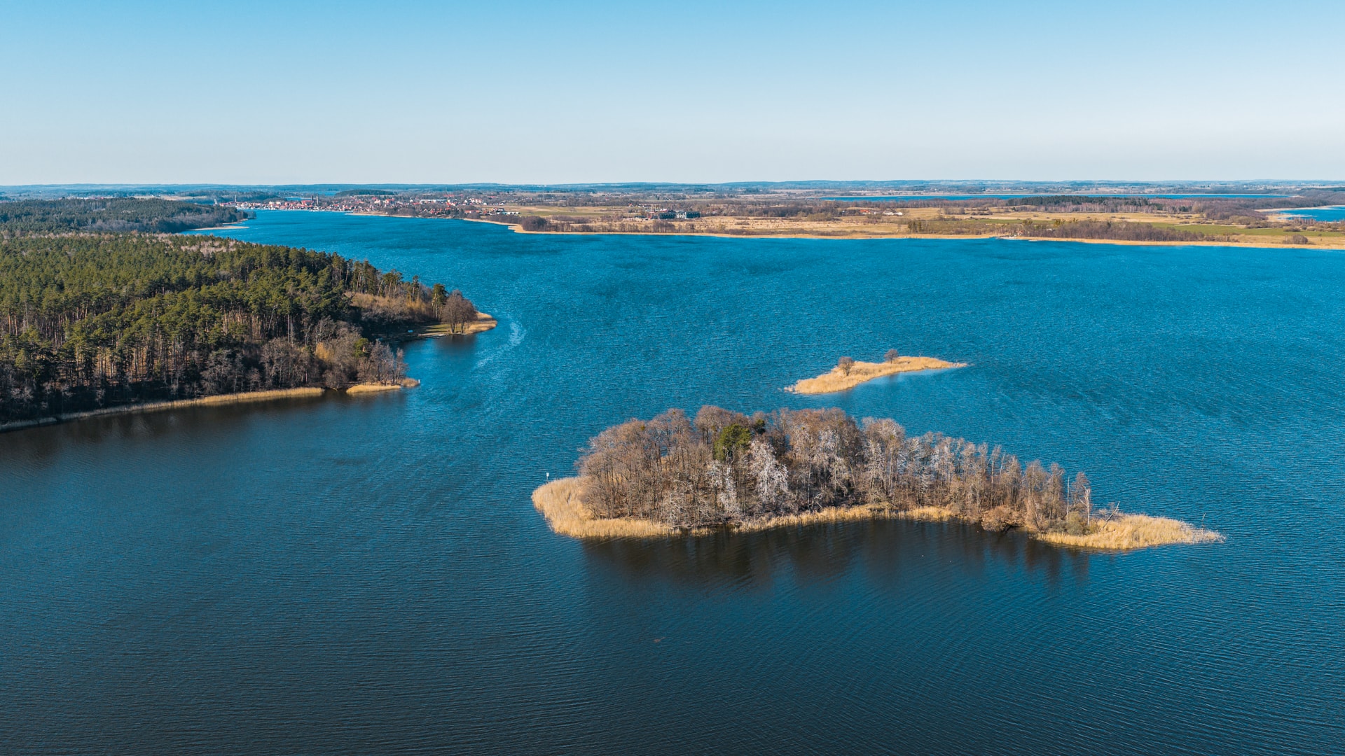 Panorama jeziora z zalesionymi wyspami, trzcinowiskami i zabudowaniami na odległym brzegu
