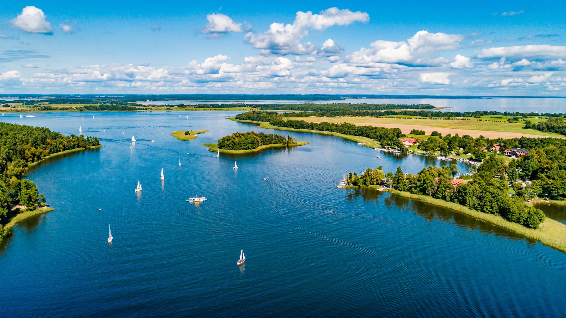 Panoramiczny widok jeziora z wyspami, żaglówkami, lasami, polami i zabudowaniami pod błękitnym niebem