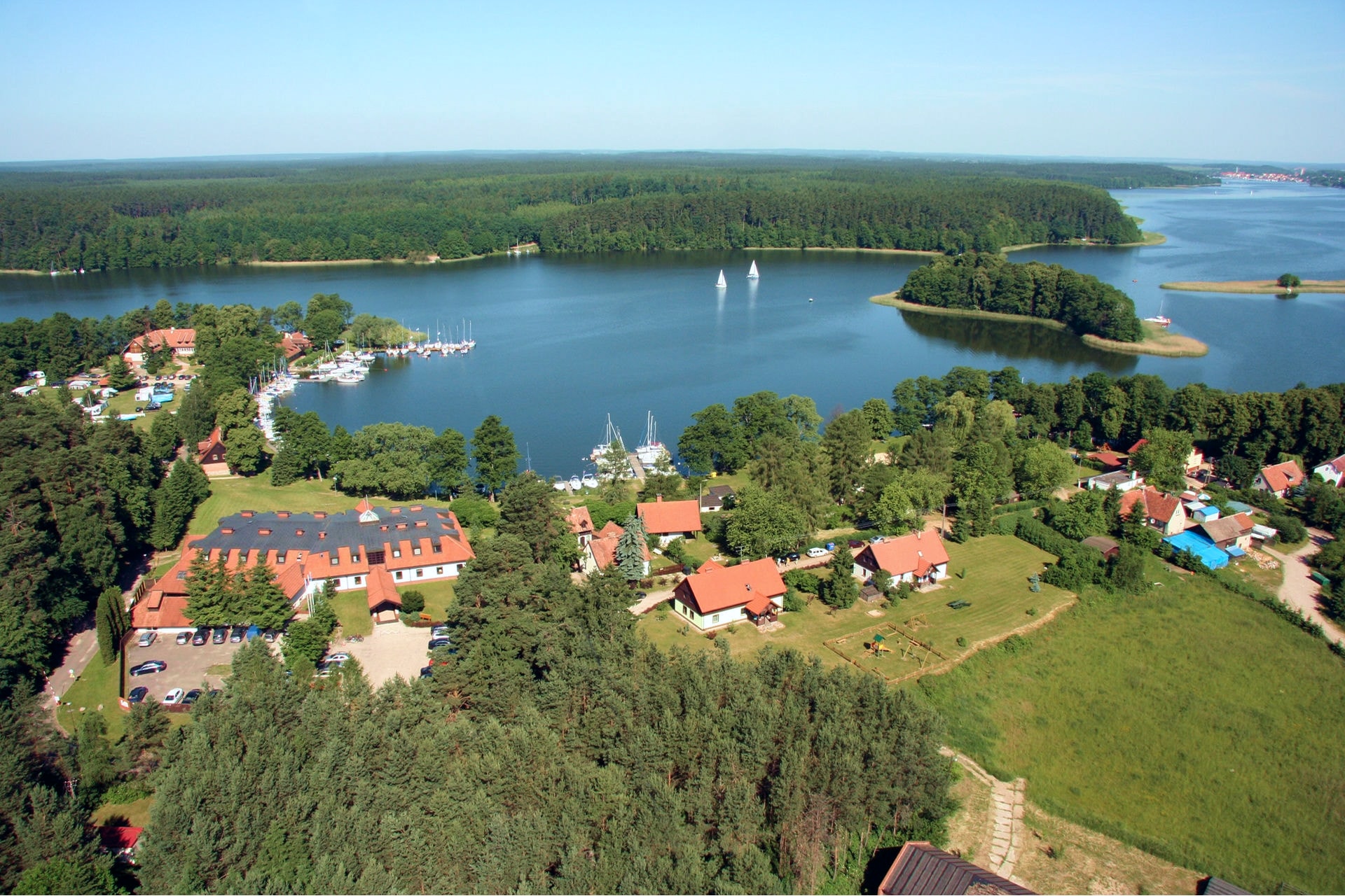 Panorama jeziora otoczonego lasami, z żaglówkami, przystanią i zabudową miejscowości na pierwszym planie