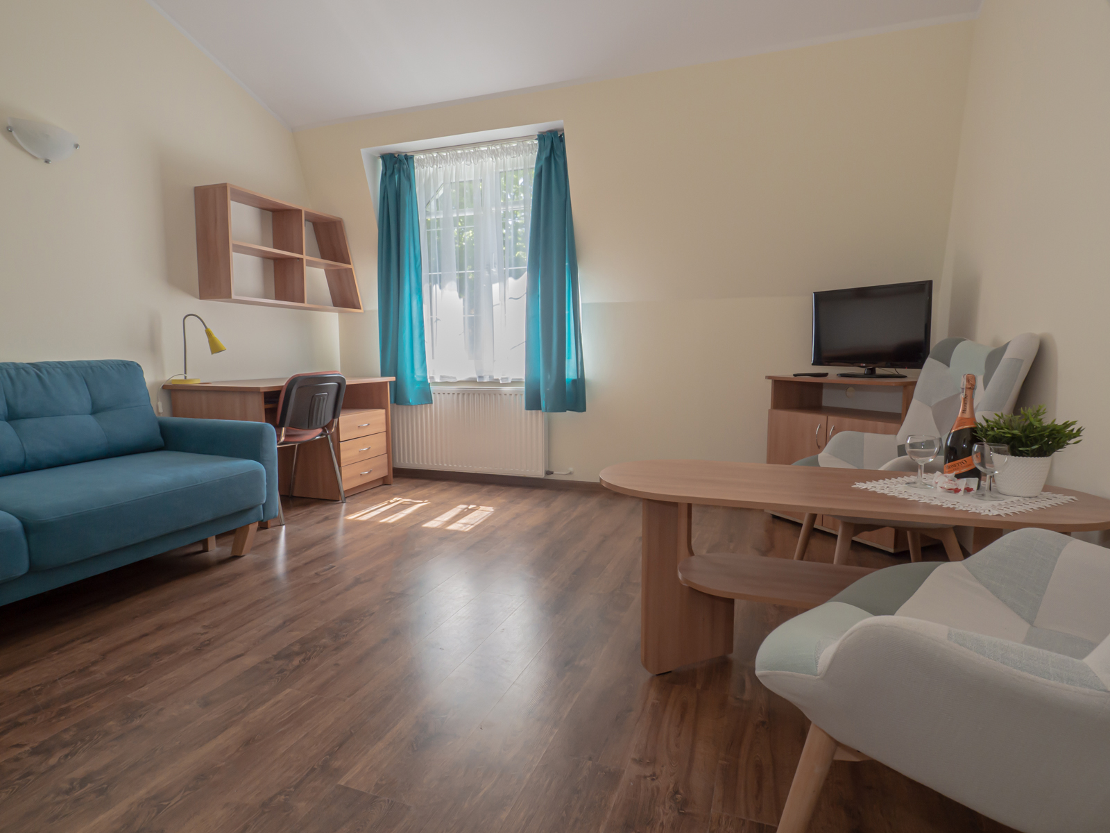 Jasny salon z niebieską sofą, biurkiem, telewizorem, stolikiem z winem i fotelami