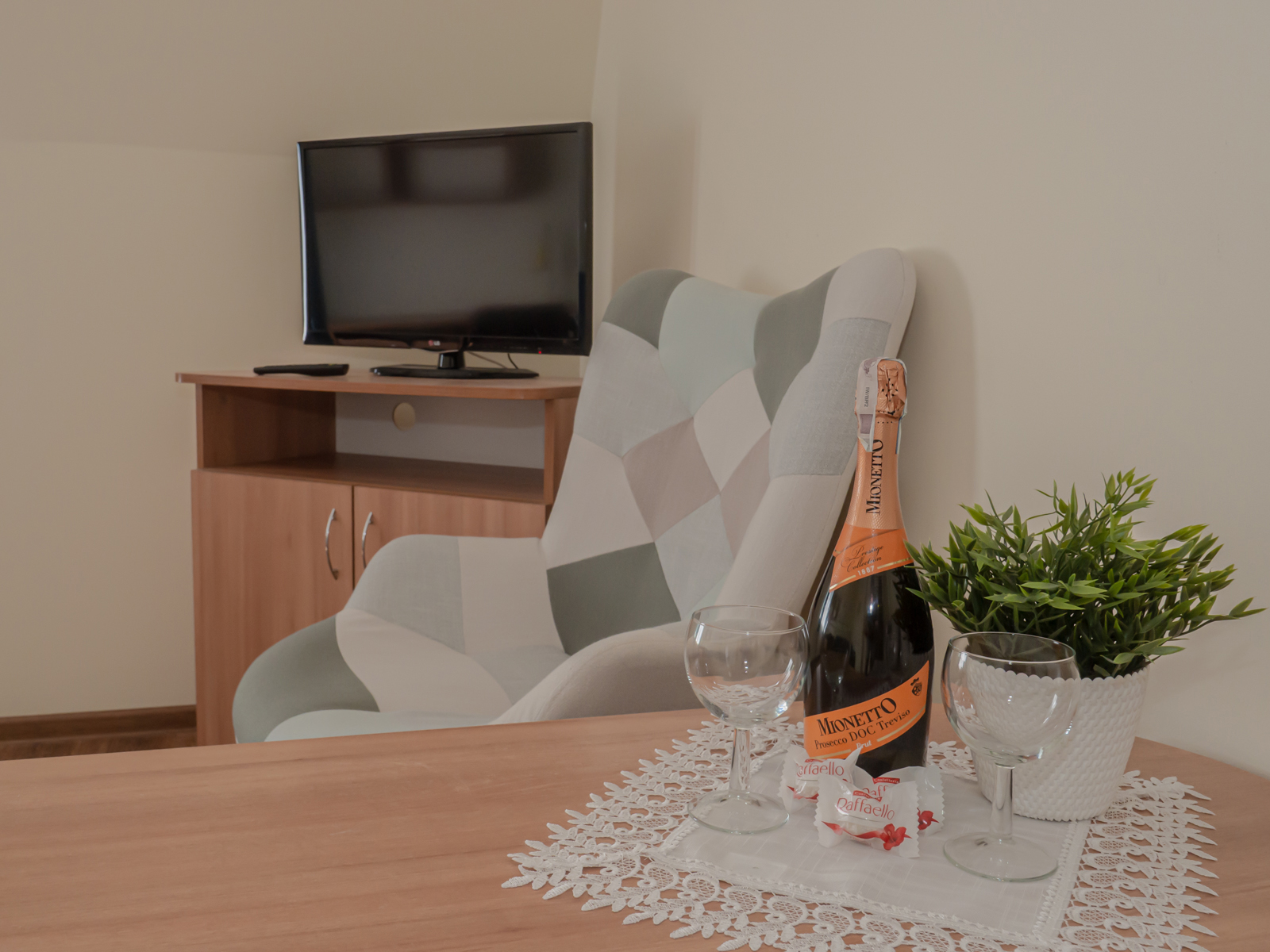 Stół z butelką prosecco, dwoma kieliszkami, pralinkami i rośliną; w tle fotel oraz telewizor