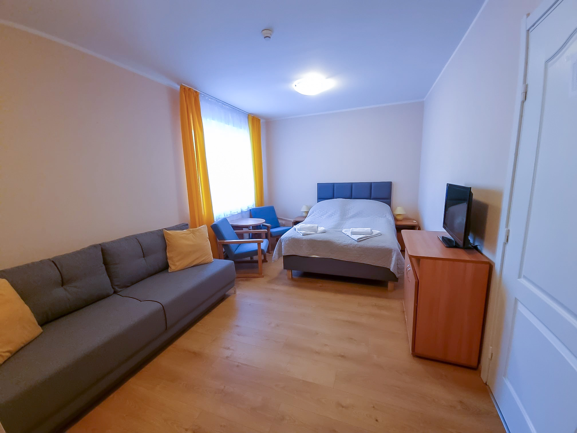 Pokój hotelowy z łóżkiem, szarą sofą, telewizorem i fotelem przy oknie z żółtymi zasłonami