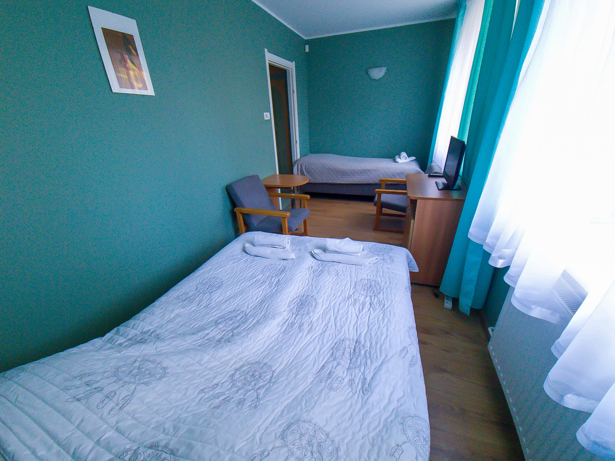 Pokój hotelowy z dwoma łóżkami, fotelami, telewizorem i dużym oknem zasłoniętym białymi firankami