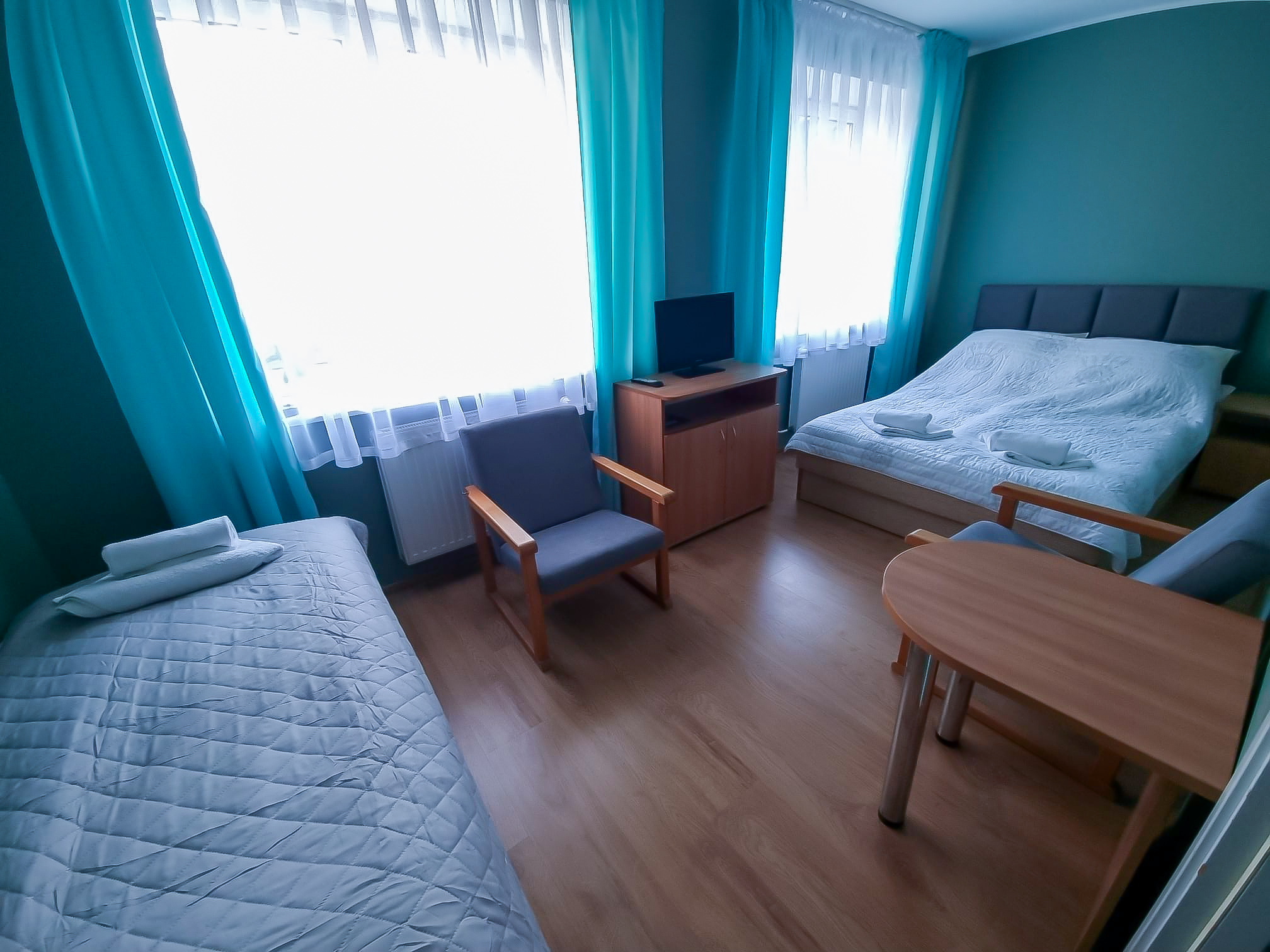 Pokój z dwoma łóżkami, telewizorem, fotelami i stołem, utrzymany w turkusowej kolorystyce