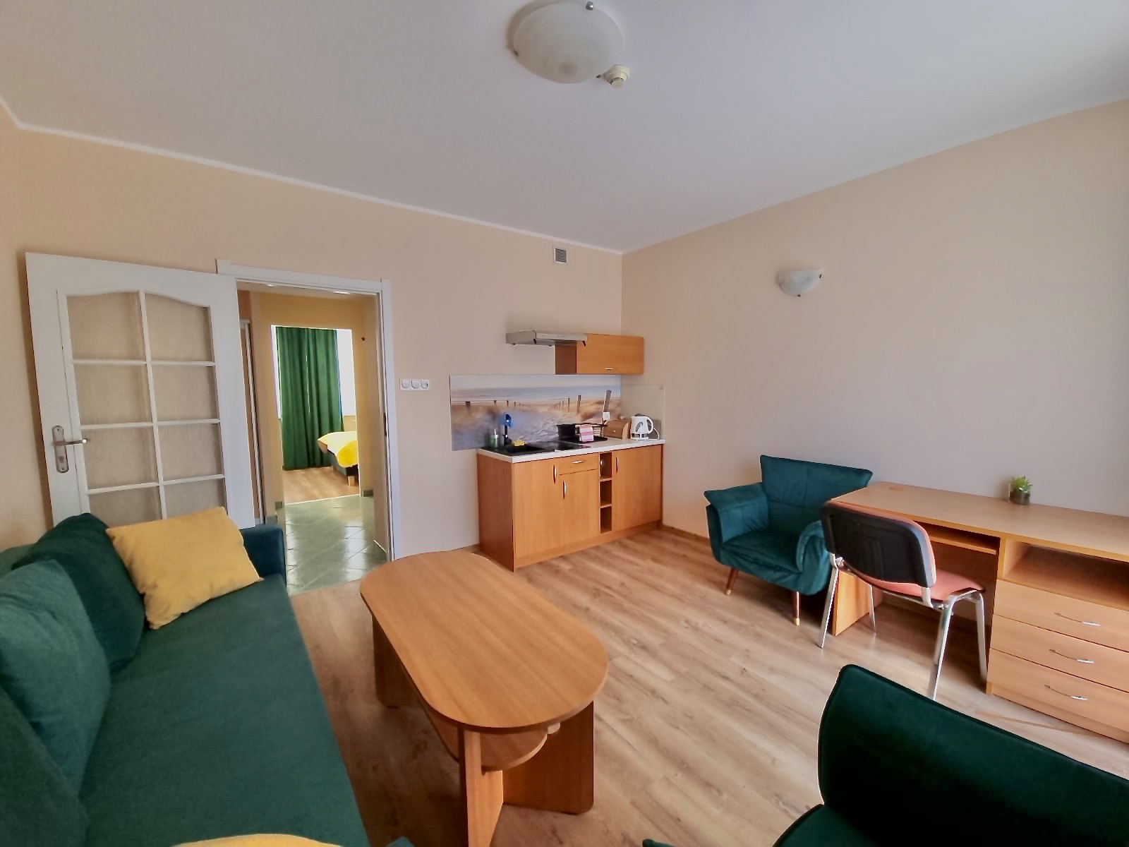 Jasny salon z aneksem kuchennym, zieloną sofą, fotelami, stolikiem kawowym i biurkiem z krzesłem