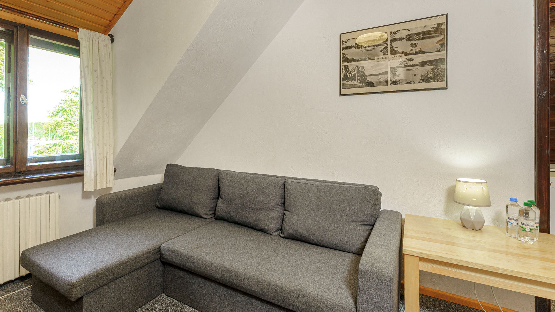 Szara narożna sofa, drewniany stolik z lampą i butelkami w jasnym pokoju pod skosem dachu