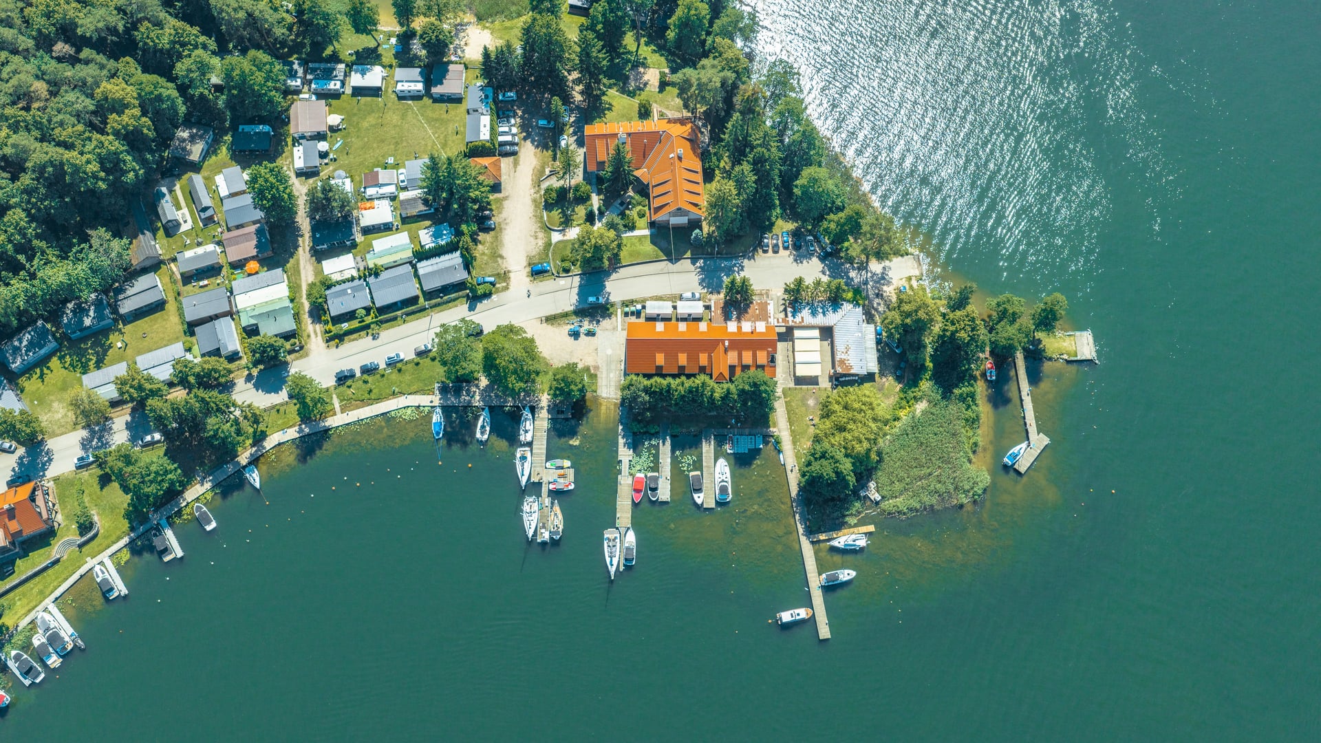 Widok z lotu ptaka na przystań nad jeziorem, domy, kemping, pomosty i liczne łodzie