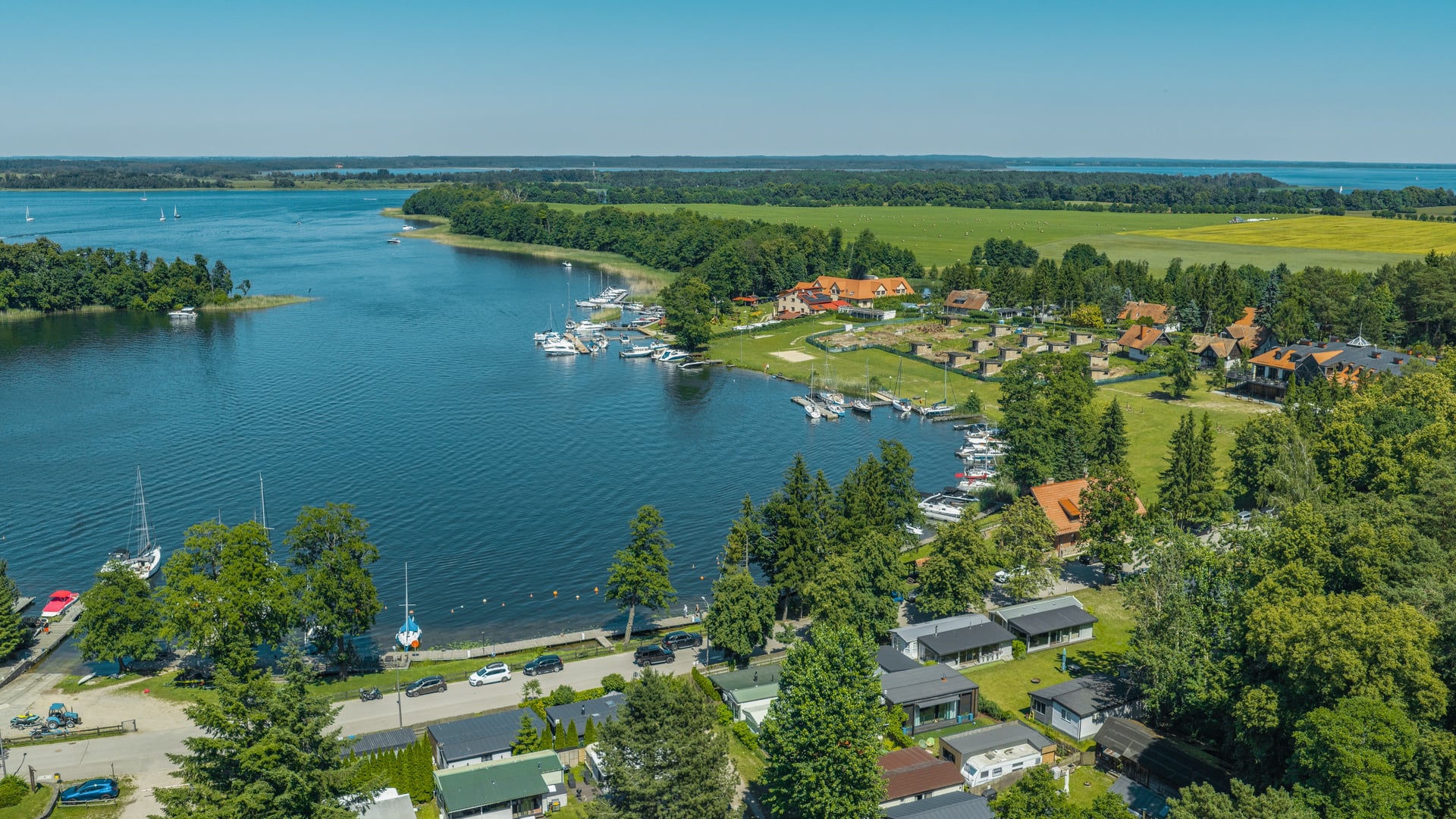 Widok z lotu ptaka na jezioro z mariną, żaglówkami, zabudową wiejską i polami uprawnymi