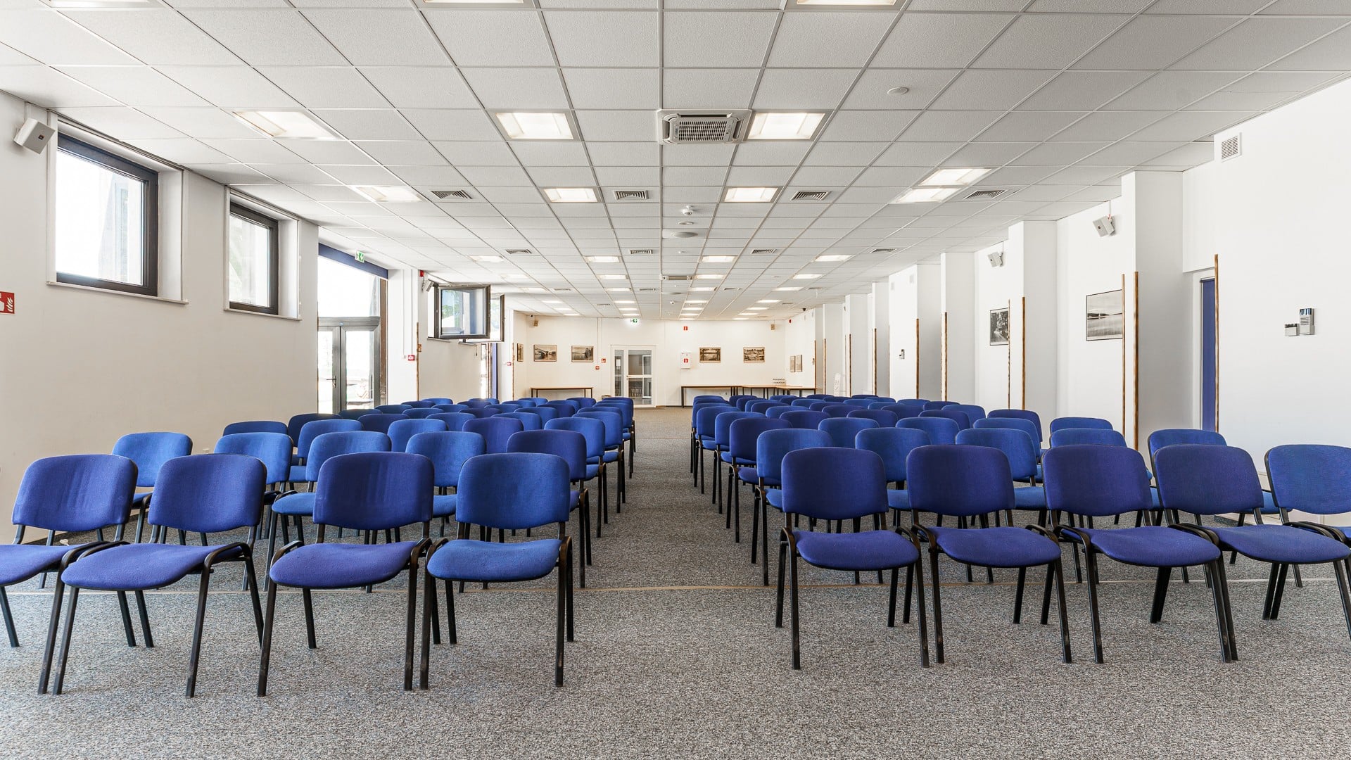 Jasna sala konferencyjna z rzędami niebieskich krzeseł i centralnym przejściem