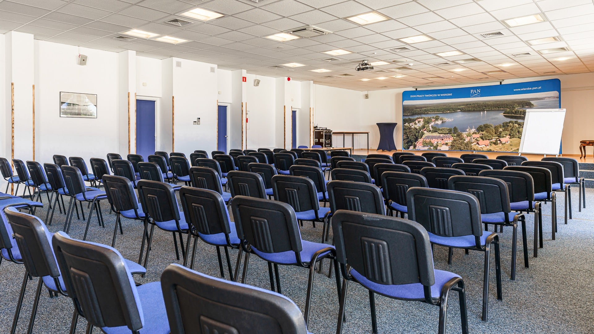 Sala konferencyjna z rzędami czarnych krzeseł, projektorem, mównicą i panoramą jeziora na ekranie