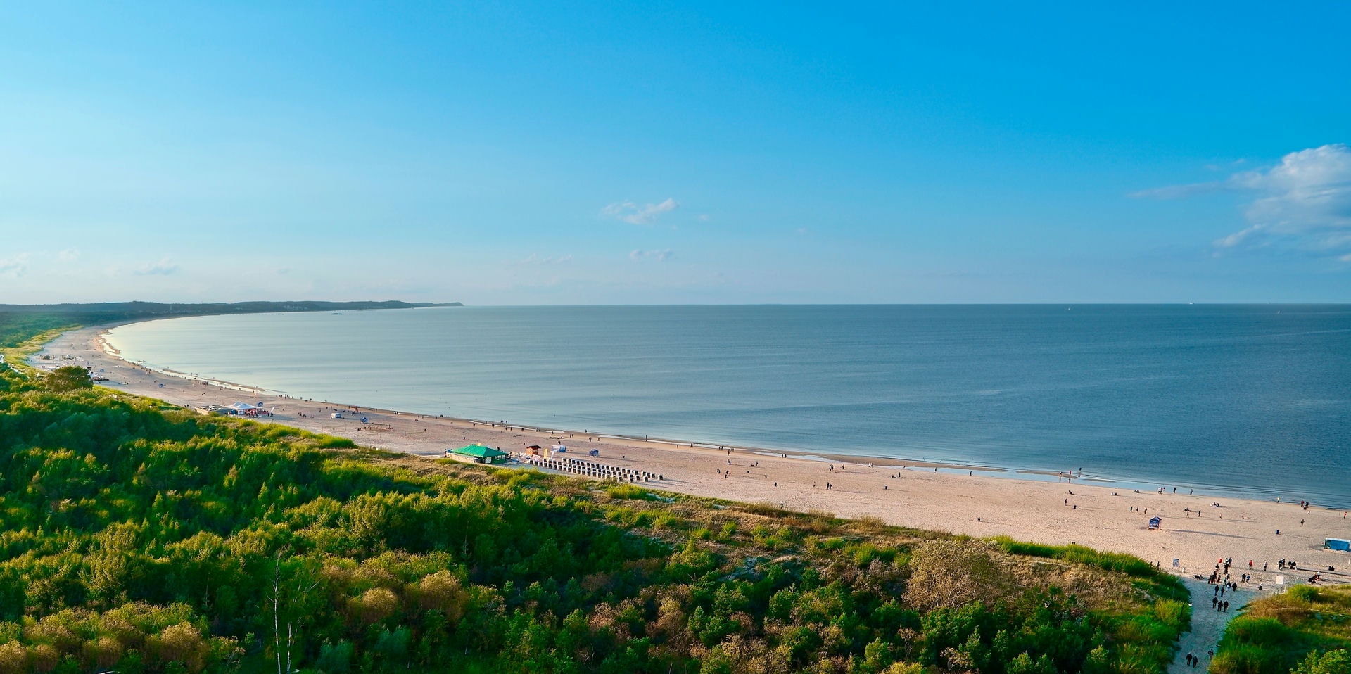 Panoramiczny widok piaszczystej plaży nad spokojnym morzem, otoczonej zielonymi wydmami i lasem